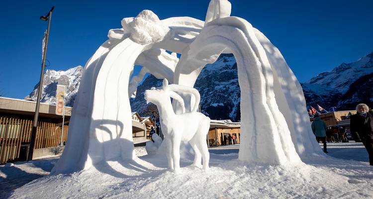 Une arche sculpturale de neige avec des figures de cerfs exposée sur une place de village alpin ensoleillée.