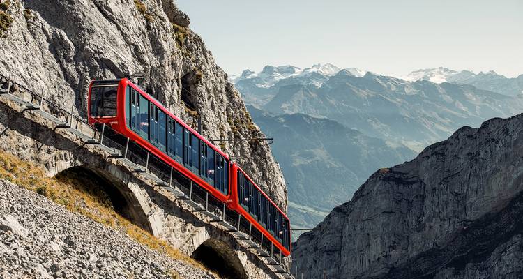 Un train à crémaillère rouge vif gravit une paroi rocheuse escarpée avec des sommets alpins panoramiques au-delà.