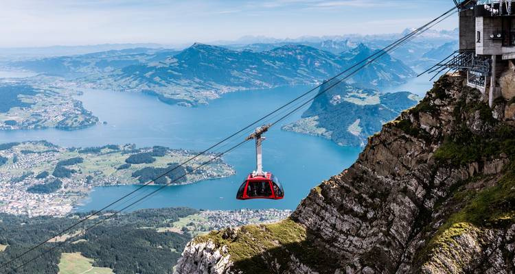 Un téléphérique rouge descend vers un vaste lac bleu et une mosaïque de villages suisses loin en contrebas.