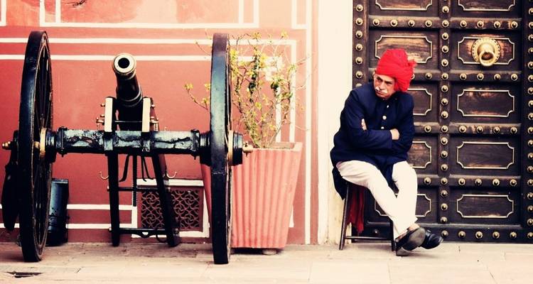 Mann mit rotem Turban sitzt mit verschränkten Armen neben einer alten Kanone vor einer verzierten rosa Palastwand und messingbeschlagenen Tür.