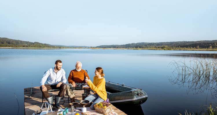 Junge Erwachsene und ein älterer Mann genießen ein Picknick auf einem hölzernen Steg an einem ruhigen See.