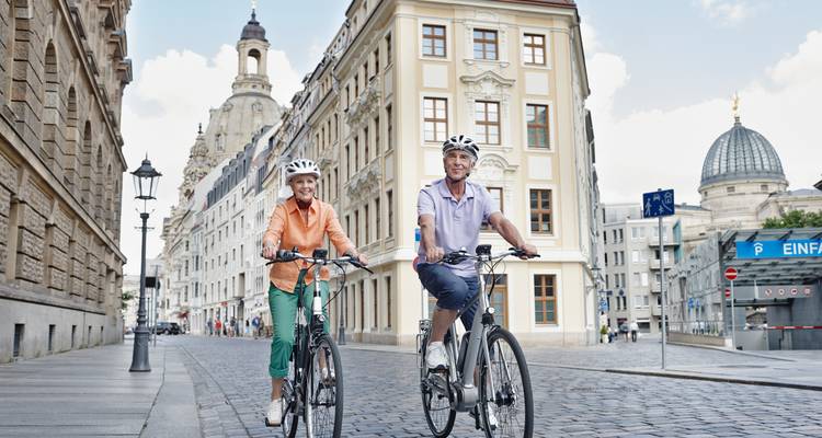 Lächelndes älteres Paar radelt durch eine historische europäische Stadtstraße, die von prächtigen Gebäuden gesäumt ist.