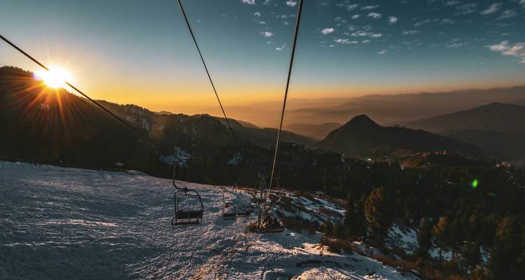 Câbles de télésiège s'étendant vers un coucher de soleil doré au-dessus de pentes enneigées et de chaînes de montagnes étagées.