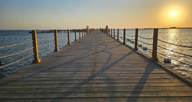 Longue jetée en bois s'étendant dans la mer Rouge au coucher du soleil avec des reflets dorés sur l'eau.