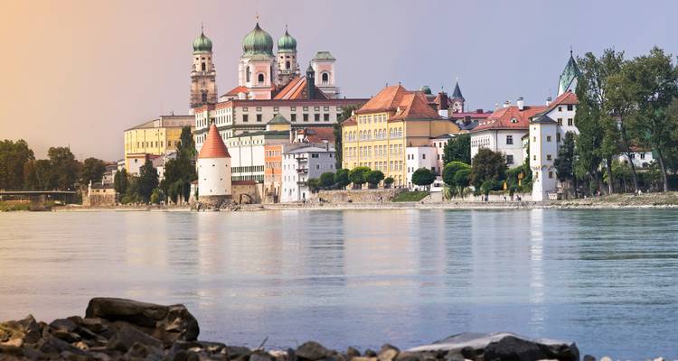 Vista pintoresca junto al río de Passau con su catedral de cúpula verde y edificios coloridos reflejándose en el Danubio durante la hora dorada.