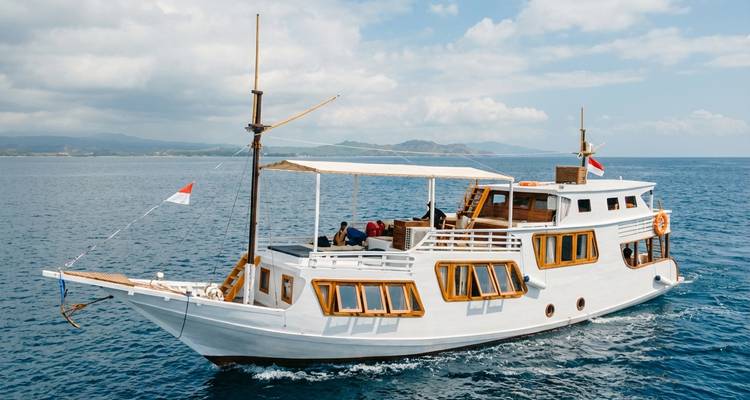 Un charmant bateau en bois habitable avec des drapeaux indonésiens navigue sur une mer bleue calme avec en arrière-plan des collines vertes lointaines et des nuages épars.