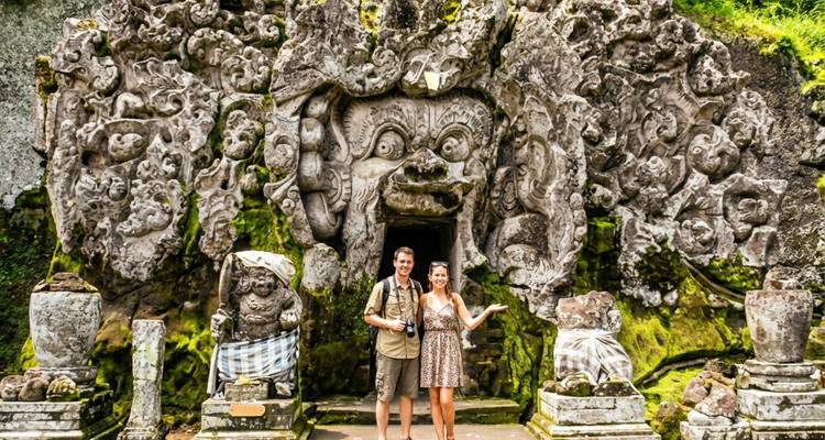 Un couple pose devant l'entrée en pierre sculptée couverte de mousse du temple Goa Gajah au milieu d'une végétation luxuriante.