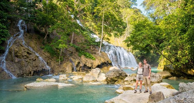 Deux randonneurs se tiennent sur des rochers à côté de bassins turquoise et d'une cascade tropicale à plusieurs niveaux entourée d'une forêt verte dense.