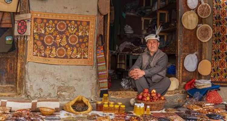 Vendedor del mercado con gorra tradicional sentado entre canastas de frutos secos, miel y artesanías fuera de una tienda rústica.