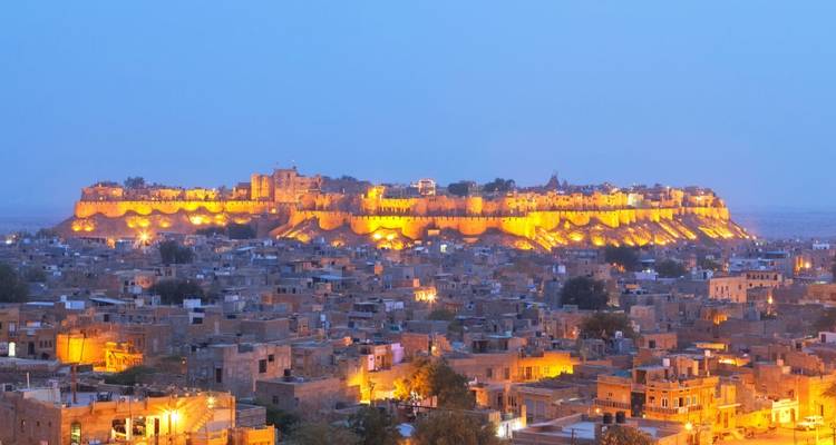 Panoramablick bei Abenddämmerung auf die beleuchtete Sandstein-Festung Jaisalmer, die sich über die flachen Dächer der Stadt erhebt.