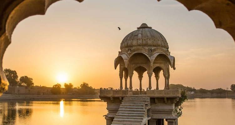 Gewölbter Blick über den Gadisar-See bei Sonnenaufgang mit einem gewölbten Sandsteinpavillon, der sich in ruhigem Wasser spiegelt.