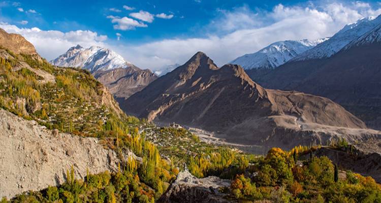 Valle expansivo con picos nevados, campos en terrazas y árboles otoñales bajo un cielo parcialmente nublado