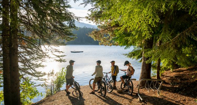 Gruppe von Radfahrern, die neben einem spiegelnden Waldsee ruhen, während das späte Nachmittagslicht auf dem Wasser funkelt.