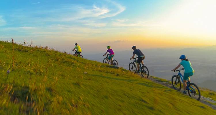 Reihe von Mountainbikern, die einen grasbewachsenen Bergrücken vor einem farbenfrohen Abendhimmel und einem entfernten Tal hinabfahren.
