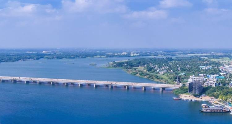 Eine lange Brücke überspannt einen breiten blauen Fluss mit grüner Landschaft und verstreuten Gebäuden dahinter.