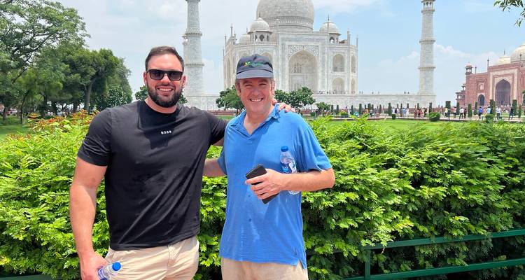 Deux voyageurs souriants devant des jardins luxuriants avec le Taj Mahal qui se dresse majestueusement derrière eux.