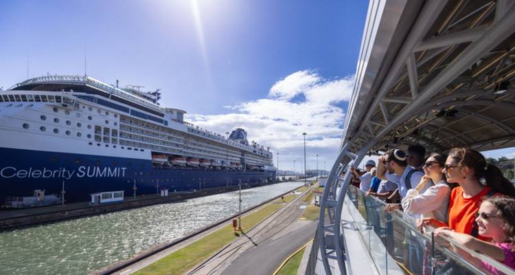 Turistas en una plataforma de observación elevada viendo un gran crucero transitar por un canal bajo un sol brillante.