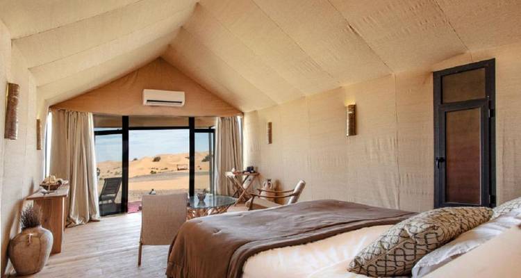 Gemütliches Wüstenlodge-Zimmer mit neutralen Farbtönen, großem Bett und Fenster mit Blick auf offene Dünen.