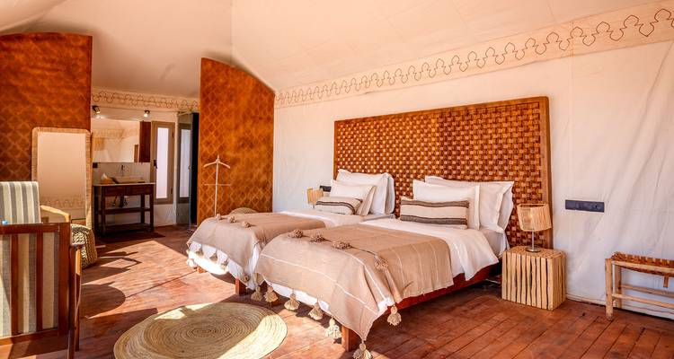 Helder tweepersoonsslaapkamertentinterieur in de woestijn met geweven hoofdeinde, houten vloeren en rustieke decoratie.