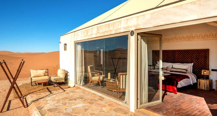 Luxe woestijnsuite met glazen wand die uitkijkt op eindeloze zandduinen onder een heldere blauwe hemel.