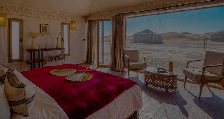 Zwak verlichte tentenslaapkamer met rode deken en woestijnuitzicht door groot raam.