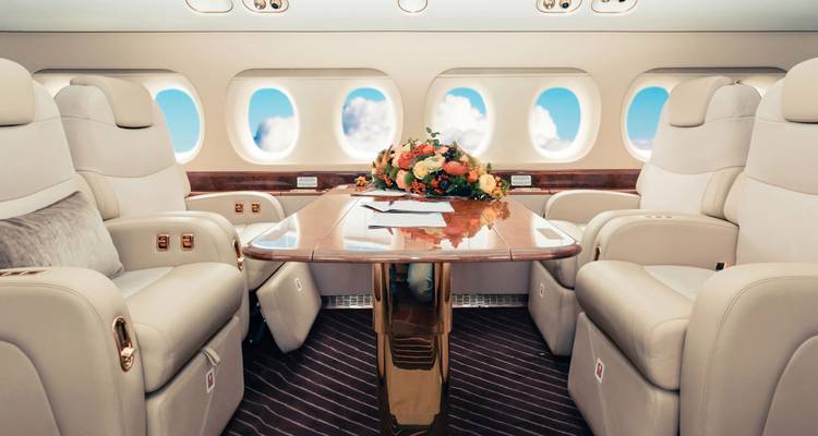 Weelderige privéjet cabine met crèmekleurige leren stoelen en bloemstuk tegen ramen met blauwe lucht.