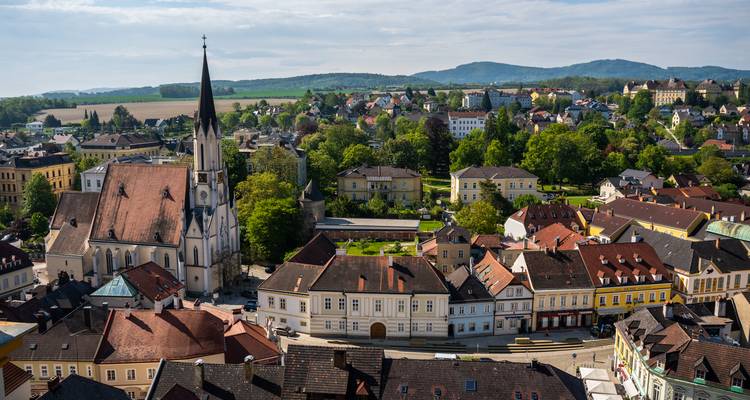 Malerische österreichische Stadt mit Kirchturm und roten Dächern inmitten sanfter grüner Hügel.