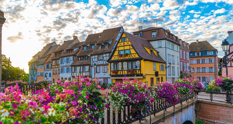 Des maisons à colombages colorées bordent un pont-canal orné de jardinières fleuries éclatantes sous un ciel dramatique.