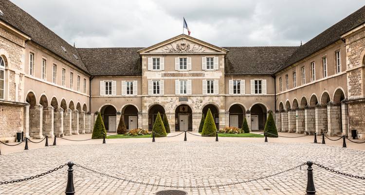 Cour symétrique d'un palais historique de Dijon avec des colonnades à arcades entourant une place pavée sous un ciel nuageux.