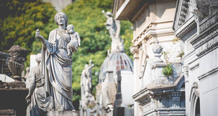 Marmor-Statuen und aufwendige Mausoleen auf einem atmosphärischen Friedhof mit Grünflächen im Hintergrund.