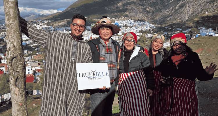 Voyageurs en tenue traditionnelle tenant une pancarte True Blue avec Chefchaouen dans la vallée en contrebas.