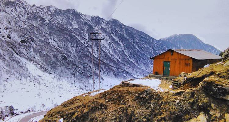 Kleine orangefarbene Hütte, die auf einem felsigen Berghang thront, umgeben von schneebedeckten Himalaya-Bergen.
