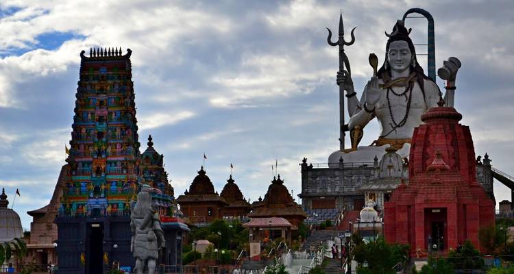 Gigantische Shiva-Statue, die über einen Komplex aus lebhaft bemalten hinduistischen Tempeln unter einem dramatischen Himmel emporragt.