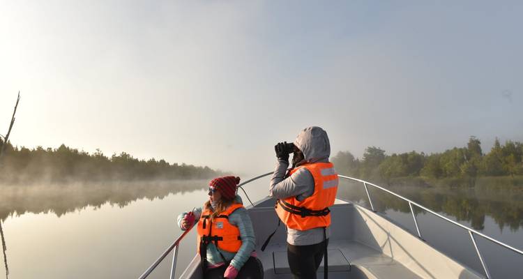 Twee reizigers in reddingsvesten observeren vogels vanuit een kleine boot op mistige, stille wateren bij dageraad.
