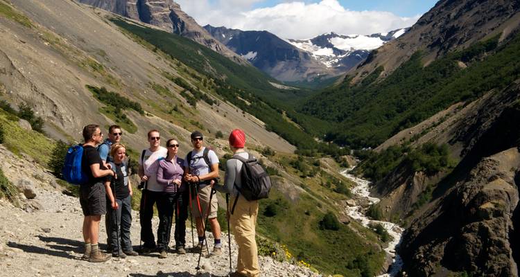Groupe de randonnée guidé fait une pause sur sentier de montagne rocheux surplombant vallée glaciaire et rivière.