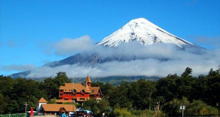 Der schneebedeckte Vulkan Osorno erhebt sich über eine bewaldete Landschaft und eine Holzhütte.