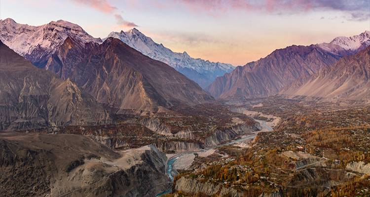 Sweeping aerial view of Hunza Valley with a turquoise river winding through rugged, snow-capped mountains at sunset.