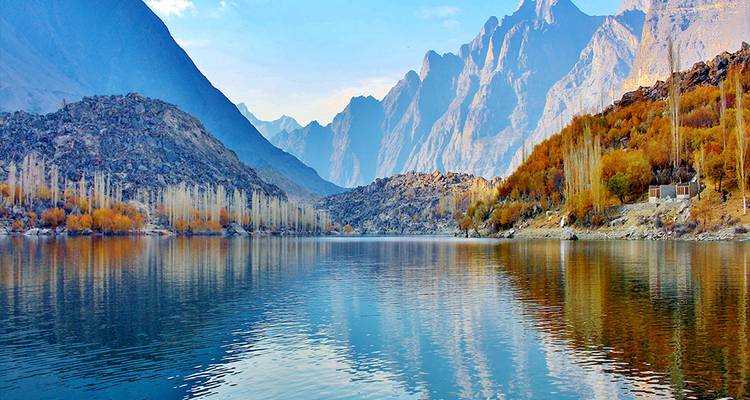 Crystal-clear alpine lake reflecting autumn trees and jagged peaks under a blue sky.