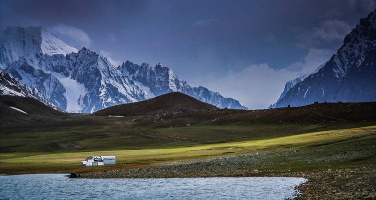 High-altitude grassland dotted with tents beside a tranquil lake, dramatic snowy peaks in the background.