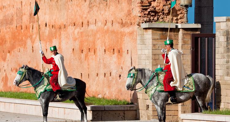 Guardias ceremoniales con uniformes rojos montan caballos grises decorados junto a las murallas ocres del complejo palaciego de Rabat.