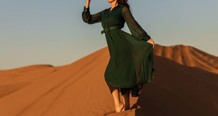 Mujer con vestido verde fluido posando descalza sobre duna de arena naranja bajo suave luz del atardecer