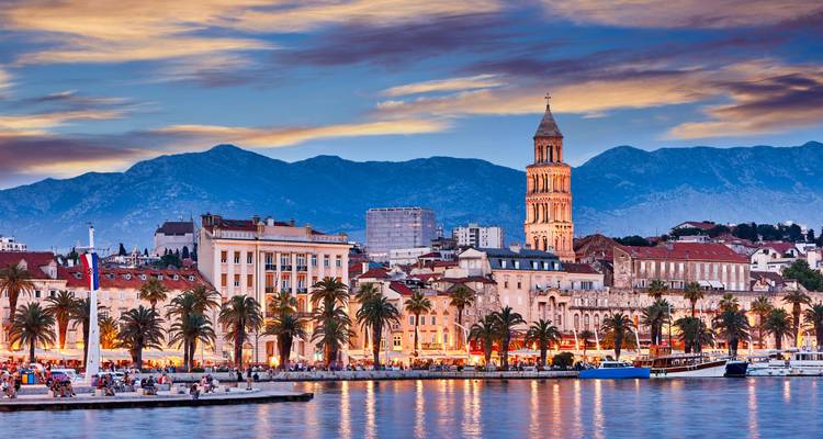 Avondverlichting van Split's boulevard aan zee met de kathedraal klokkentoren die oprijst tegen de schemering bergen.