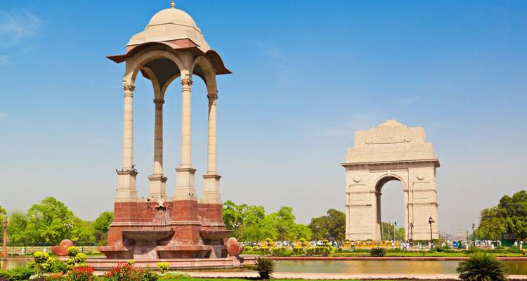 Helder blauwe lucht zicht op New Delhi's India Gate gedenkteken en nabijgelegen bladerdak weerspiegeld in een vijver.