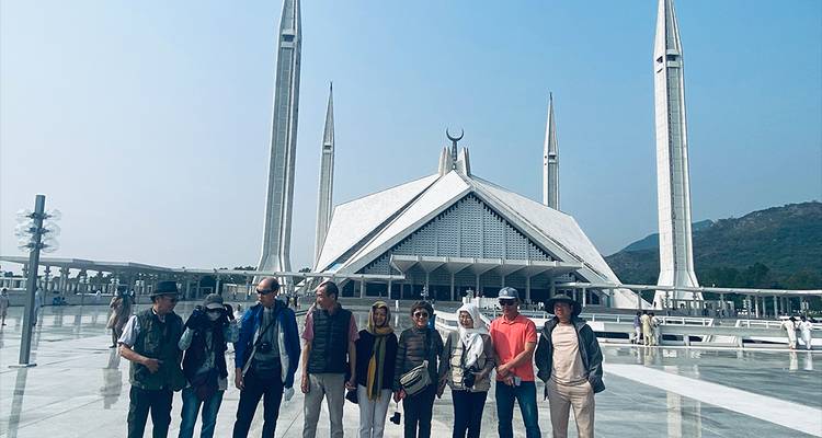 Un grupo de turistas posa en la plaza reluciente frente a la icónica Mezquita Faisal de Islamabad bajo un cielo azul despejado.