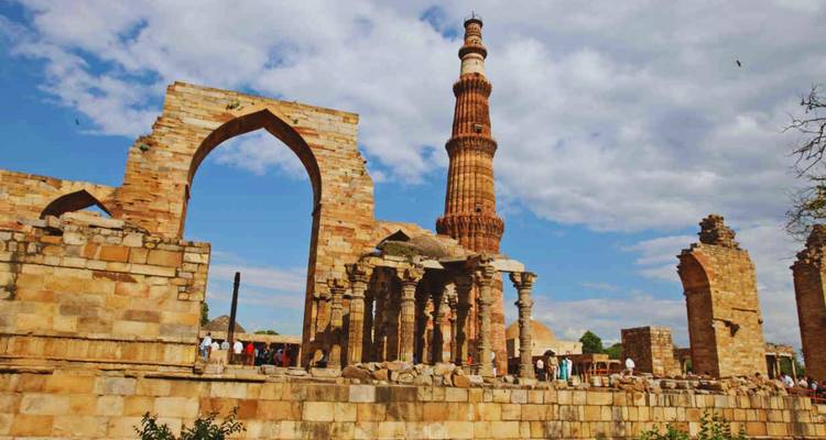 Stenen booggewelven en de torenhoge Qutub Minar rijzen op onder een gedeeltelijk bewolkte hemel.