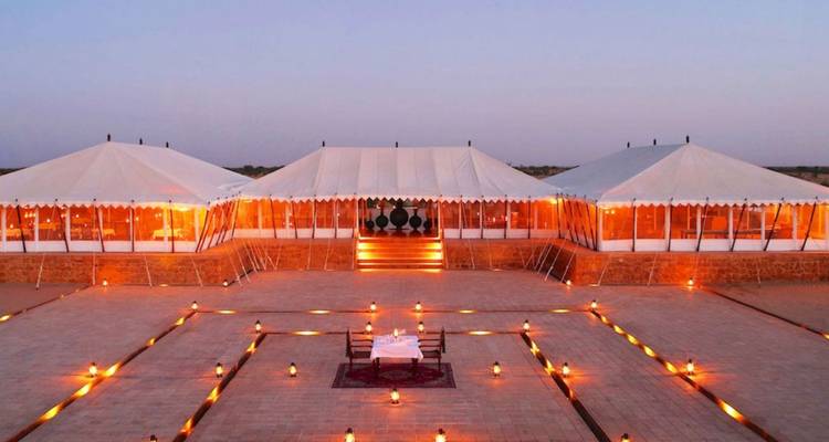 Campement de luxe dans le désert avec des tentes en toile blanche éclairées chaleureusement au crépuscule par des lanternes qui bordent la cour.