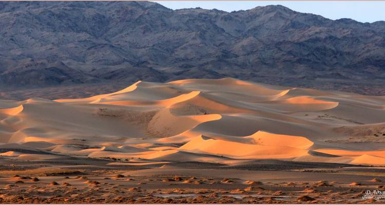 Des dunes de sable ondulantes teintées de lumière chaude avec des montagnes escarpées en arrière-plan au loin.