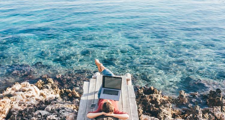 Eine Person lehnt sich auf einem Holzsteg über kristallklarem türkisfarbenem Wasser zurück, arbeitet an einem Laptop und genießt die ruhige Meereslandschaft.