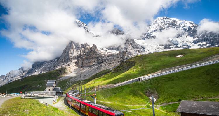 Roter Bergzug fährt grüne Alpenhänge hinauf in Richtung schneebedeckter Gipfel unter vereinzelten Wolken.