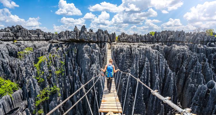 Routard traversant un pont suspendu étroit au-dessus de formations calcaires spectaculaires aux pointes acérées sous un ciel lumineux.